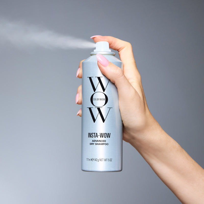 Hand holding silver Color Wow Insta-Wow Advanced Dry Shampoo spray can, 171ml / 142g / NET WT 5.0oz, releasing mist against a neutral gray background.