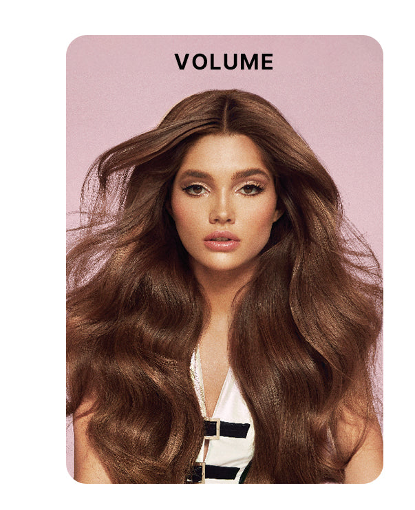 Model with long, full-bodied, voluminous brunette hair styled in loose waves, wearing a white top with black accents against a soft pink background. Bold black text above reads: “VOLUME.”