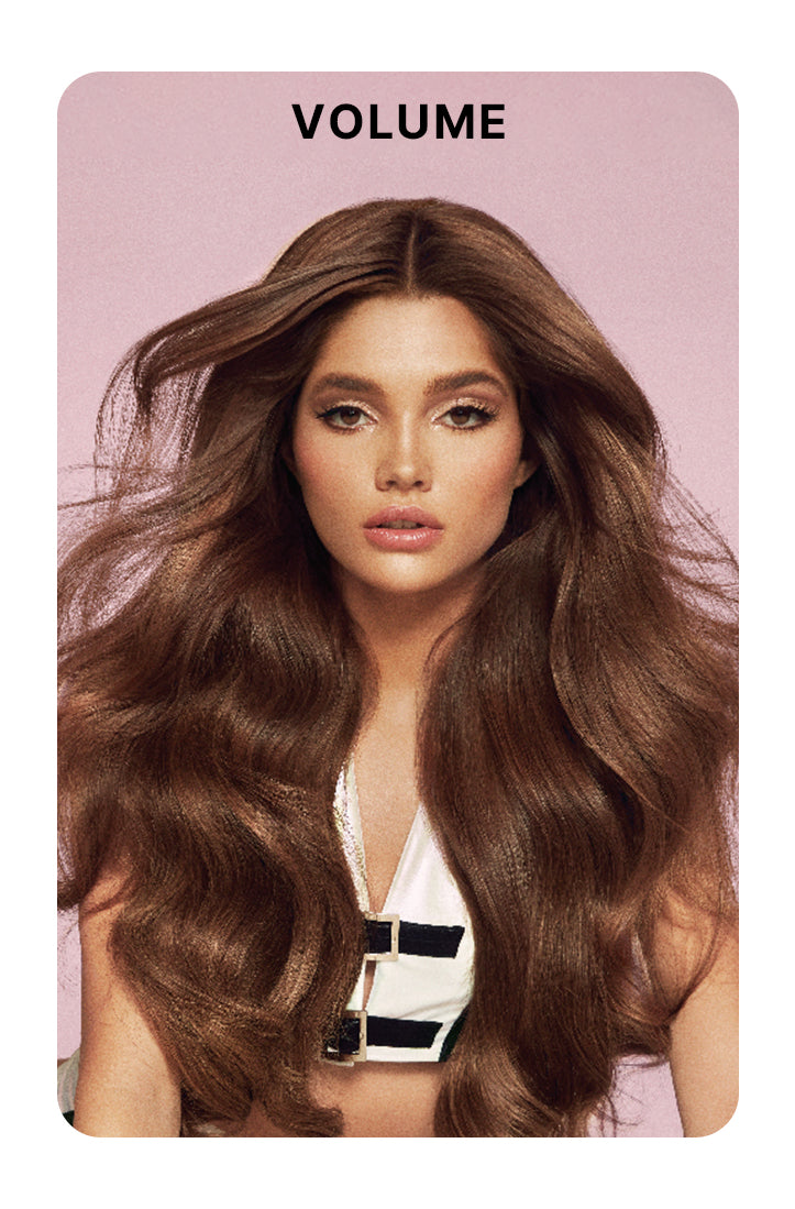 Model with long, full-bodied, voluminous brunette hair styled in loose waves, wearing a white top with black accents against a soft pink background. Bold black text above reads: “VOLUME.”